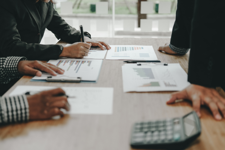 Financial advisors reviewing charts and data during a meeting, with hands pointing and writing on documents that show financial graphs and charts.